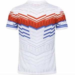 May include: White short-sleeved shirt with a geometric pattern. The shirt features red and blue accents across the shoulders and sleeves. The design includes chevron and floral elements, creating a visually striking pattern.