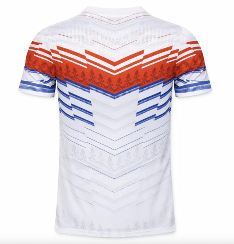 May include: White short-sleeved shirt with a geometric pattern. The shirt features red and blue accents across the shoulders and sleeves. The design includes chevron and floral elements, creating a visually striking pattern.