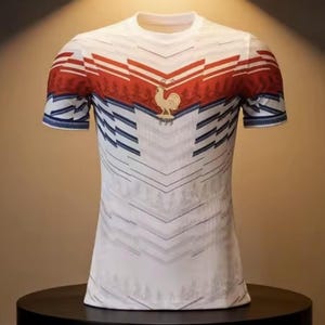 May include: White t-shirt with a red and blue design. The shirt features a red band across the chest and shoulders, with a gold rooster emblem. Blue and white diagonal lines create a pattern on the front and sleeves. The shirt has a crew neck and short sleeves.