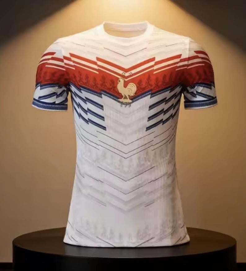 May include: White t-shirt with a red and blue design. The shirt features a red band across the chest and shoulders, with a gold rooster emblem. Blue and white diagonal lines create a pattern on the front and sleeves. The shirt has a crew neck and short sleeves.