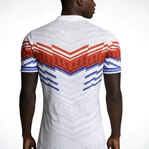 May include: White polo shirt with a geometric design in red, blue, and white. The shirt features a collar and short sleeves. The back of the shirt has a chevron pattern with floral accents.