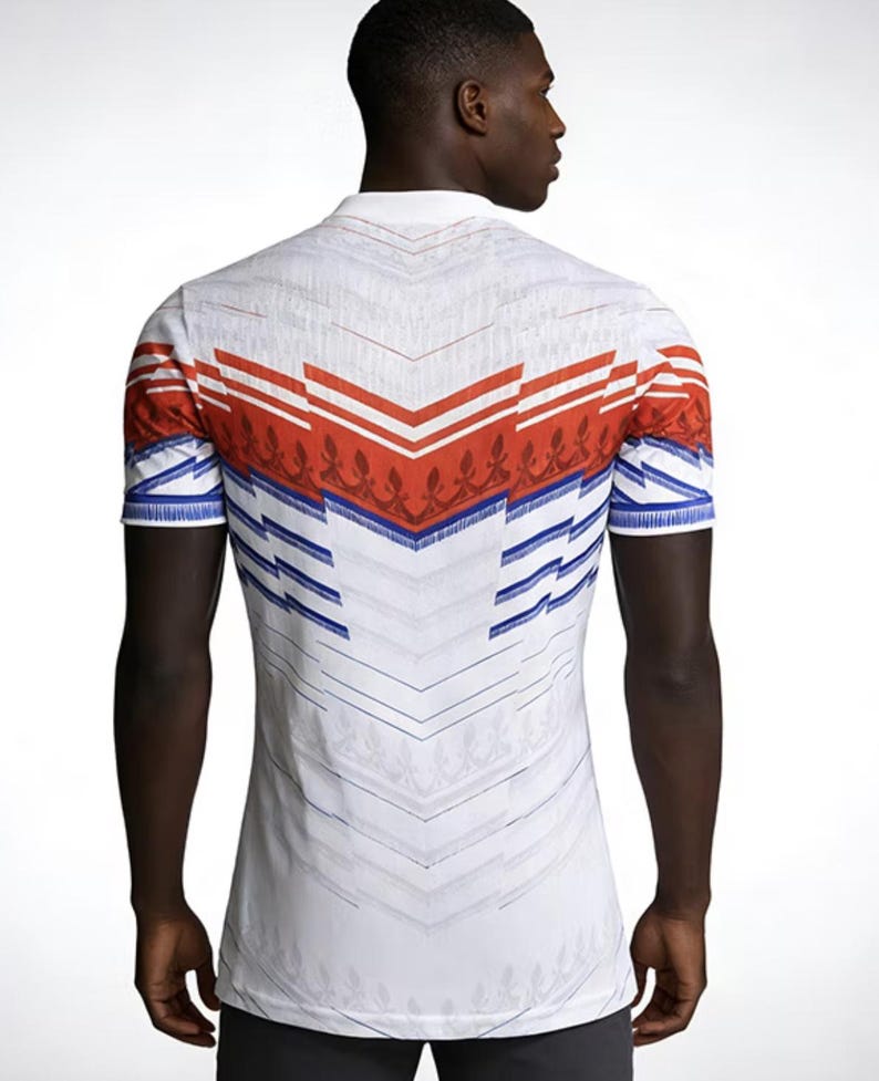May include: White polo shirt with a geometric design in red, blue, and white. The shirt features a collar and short sleeves. The back of the shirt has a chevron pattern with floral accents.