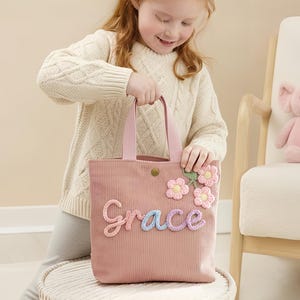 May include: A pink corduroy tote bag with the name "Grace" in colourful script. The bag features floral embellishments and a button closure. A young girl holds the bag, with a pink elephant plush toy and a chair in the background.