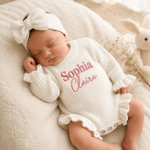 Könnte beinhalten: Ein cremefarbener, langärmeliger Strickstrampler mit gerüschten Kanten und dem in rosa Garn auf die Brust gestickten Namen Sophia Claire, kombiniert mit einem passenden Strickstirnband mit einer großen Schleife am Oberkopf.