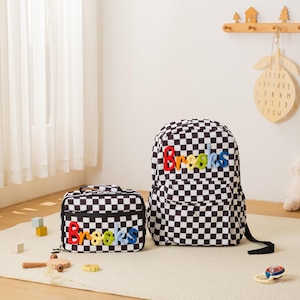 May include: A black and white checkered backpack and matching lunch bag. Both items have the name "Brooks" embroidered in colorful letters. The backpack has a black shoulder strap.