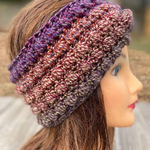 May include: A wide, crocheted headband featuring a variegated yarn pattern in shades of purple, orange, and earthy brown. The accessory has a textured, ribbed stitch design that wraps around the head, showcasing a thick, warm construction.