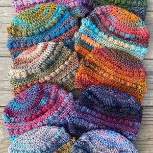 May include: A collection of colorful, hand-knitted beanies. Each hat features a textured, bobble stitch design and is made with a variety of vibrant yarn colors, including pink, blue, green, and orange. The hats are displayed on a wooden surface.