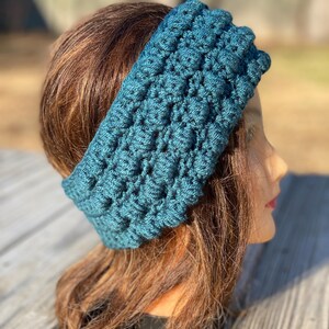 May include: A dark teal crochet headband featuring a textured bobble stitch pattern. The accessory is wide and wraps around the head, displaying a consistent, repeating raised knot design throughout the entire surface of the yarn-based material.