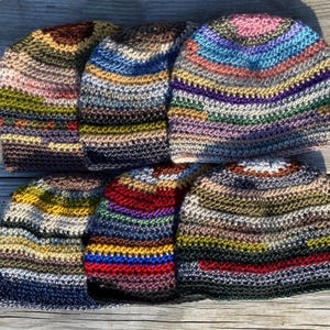May include: A collection of seven colorful, handmade crochet beanies. Each hat features a unique combination of colors, including blues, greens, reds, and yellows. The hats are arranged on a wooden surface, showcasing their textured, striped designs.