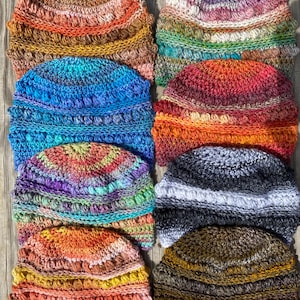 May include: A collection of handmade crochet hats in various colors. The hats feature a textured, bobble stitch design and are arranged on a wooden surface. Colors include blues, oranges, greens, and purples, with some hats displaying a gradient effect.