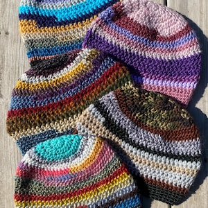 May include: A collection of five handmade crochet beanies in various colors. Each hat features horizontal stripes in a mix of colors, including blue, purple, yellow, red, and green. The hats are displayed on a wooden surface.