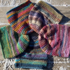 May include: Five crocheted beanies arranged in a circular pattern. Each hat features a distinct variegated yarn color scheme, including combinations of bright rainbow, muted earth tones, vibrant pinks, deep greens, and dark blues with purple accents.
