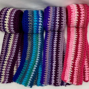 May include: Four handmade crochet scarves in shades of purple, blue, teal, and pink. Each scarf features a striped pattern with a textured stitch. The scarves are rolled up at the top, showcasing the color variations.