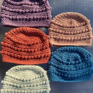 May include: A collection of five crocheted beanies in purple, tan, rust orange, light green, and dark blue. Each hat features a textured pattern of horizontal rows of raised bobble stitches alternating with smooth, flat crochet stitch bands.