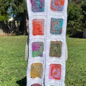 May include: A long crochet scarf featuring a series of square motifs in various variegated colors, including orange, blue, green, and purple, all bordered by thick white yarn. The scarf is displayed on a mannequin, showing a repeating pattern.