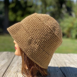 May include: A tan crochet bucket hat with a slightly angled brim. The hat is made of a textured, woven material and is displayed on a mannequin head. The background is a blurred view of green trees and a wooden surface.