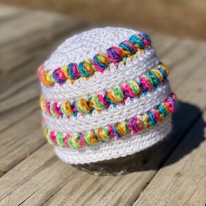 May include: A white crocheted beanie featuring three horizontal rows of colorful, variegated yarn bobbles. The bobbles incorporate bright shades of pink, orange, yellow, green, and blue, creating a vibrant contrast against the solid white base.