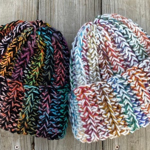 以下が含まれることがあります： 2つのニット帽のクローズアップ。1つは主に黒で虹色のアクセントがあり、もう1つは白で虹色の色合いがあります。どちらも折り返しがあり、太い糸で作られています。木製の表面に帽子が展示されています。