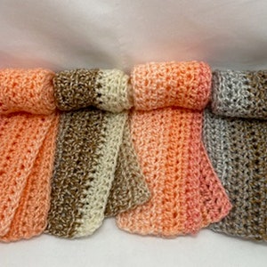 May include: Four handmade crochet scarves in shades of peach, brown, and gray. Each scarf is rolled at the top, with a textured, open weave design. The scarves are displayed on a white surface.