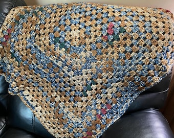 Puede incluir: Manta de ganchillo hecha a mano con un patrón de cuadrados de la abuela. La manta presenta una mezcla de hilos azules, marrones y beige, con pequeños acentos verdes y rosas. La manta está sobre una superficie negra.