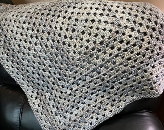 Puede incluir: Una manta hecha a mano, tejida a ganchillo con un patrón de cuadrados de la abuela. La manta presenta una mezcla de hilos grises, blancos y azules, creando un diseño texturizado y de tejido abierto. La manta está sobre una superficie negra.