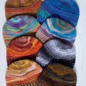 May include: A collection of handmade crochet beanies in various colors. The beanies feature a mix of warm and cool tones, including blues, oranges, purples, and browns. Each hat has a unique color pattern and texture, showcasing the craftsmanship of the crochet work.