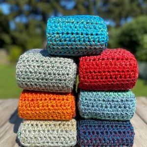 May include: A stack of nine colorful, handmade crochet cozies. The cozies are in various colors, including blue, red, orange, and green. The crochet stitch creates a textured appearance. The cozies are stacked on a wooden surface with a blurred green background.