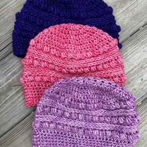 May include: Three crocheted beanies stacked vertically, featuring a textured bobble stitch pattern. The top beanie is deep purple, the middle beanie is bright pink, and the bottom beanie is a light lavender purple, all crafted with thick yarn.