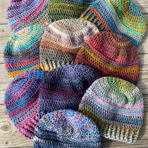 May include: A collection of colorful, handmade crochet beanies. Each hat features a unique blend of vibrant colors, including blues, pinks, greens, and purples. The beanies are displayed on a wooden surface, showcasing their textured, handcrafted details.