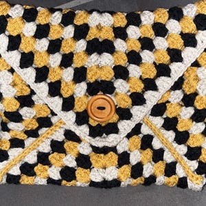 Könnte beinhalten: Eine gehäkelte Kupplungsumhängetasche mit einem geometrischen Muster in Schwarz, Weiß und Gold. Die Clutch hat einen Verschluss mit einem Holzknopf.