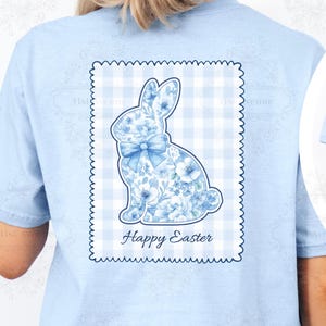 Peut inclure: T-shirt bleu clair avec un motif de lapin floral et un nœud, sur un fond à carreaux. Les mots "Happy Easter" sont imprimés sous le lapin. Un motif de lapin plus petit est sur le devant du t-shirt.