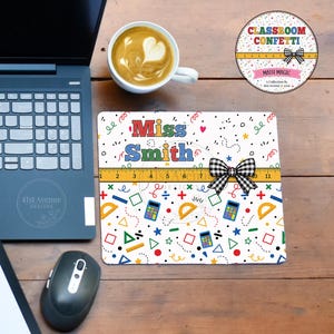 May include: A rectangular mouse pad featuring a white background with colorful math-themed icons, a yellow ruler graphic, a black and white gingham bow, and the text "Miss Smith" in multi-colored letters, positioned next to a laptop and mouse.