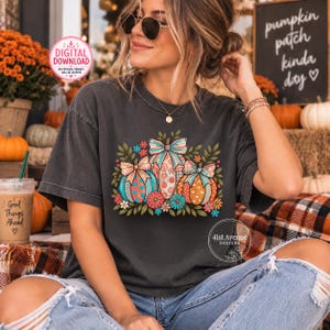May include: A charcoal gray t-shirt featuring a graphic of three pumpkins decorated with colorful polka dots, stripes, and bows, surrounded by green leaves and small flowers. The shirt has a crew neckline and short sleeves, worn with blue jeans.