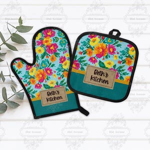 Puede incluir: Un juego de manopla y agarradera con estampado floral. El fondo turquesa está adornado con flores amarillas, naranjas y rosas. Cada pieza tiene una etiqueta marrón con el texto "Beth's Kitchen".