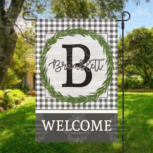 May include: A black and white checkered welcome garden flag with a green wreath and the letter B in the center. The wreath has the text "Brankslett" inside. The bottom of the flag says "Welcome".