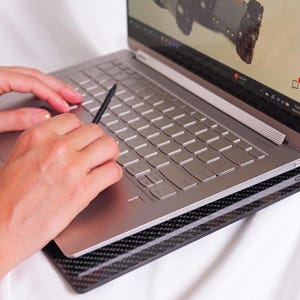 Könnte beinhalten: Ein silberner Laptop mit metallischer Tastatur und einem schwarzen Eingabestift auf dem Trackpad. Der Laptop steht auf einer schwarzen Kühlunterlage mit Carbonfaser-Muster. Der Bildschirm zeigt eine digitale Benutzeroberfläche.