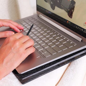 Op de afbeelding: Een zilveren laptop met een zwarte stylus op het toetsenbord. De laptop staat op een zwart-grijs patroon oppervlak. Het scherm toont een afbeelding. De laptop staat op een witte ondergrond.