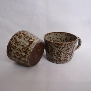 May include: Two ceramic mugs with a speckled brown and white glaze. One mug has a handle and the other does not.