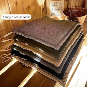 May include: A stack of square sauna pillows in various colors, including brown, olive green, and black, with white loops. The pillows are on a wooden surface, with a sign that says "Relax Steam Refresh". The text "Many color options!" is also visible.