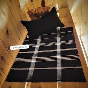 May include: A black rectangular rug with horizontal stripes in light pink and white, and a black pillow. The rug is placed on a wooden surface. The text "4 Color Options!" is in the lower left corner.