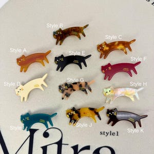 Könnte beinhalten: Eine Sammlung von katzenförmigen Haarspangen in verschiedenen Farben und Mustern, beschriftet von Style A bis Style K. Die Spangen haben gebogene Körper, spitze Ohren und kleine Schwänze, mit einfarbigen, schildpattfarbenen oder irisierenden Oberflächen.
