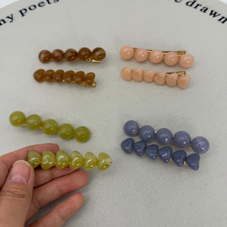 A flat lay photo showing four sets of beaded hair clips arranged on a white background.
Each set consists of two gold-tone alligator clips: one featuring a row of round spherical beads and the other featuring a row of heart-shaped beads.
The sets come in four glossy, jelly-like colors:

Toffee Brown (top left).
Peachy Pink (top right).
Matcha/Olive Green (bottom left).
Dusty Blue/Grey (bottom right).