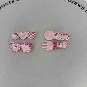 Può includere: Una collezione di fermagli e forcine rosa per capelli con disegni di Hello Kitty. I fermagli includono forme di cuore, un fiocco e un design a forchetta e cucchiaio. Alcuni fermagli hanno un motivo leopardato.