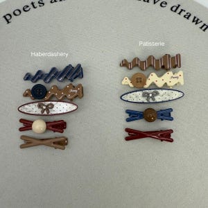May include: A collection of hair clips in various designs and colors. The clips include wavy patterns, button accents, and bow details. The color palette features shades of brown, blue, red, and white, with the word "Fancy" on some clips.