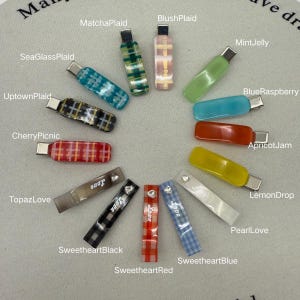 May include: A collection of colorful hair clips in various designs. The clips feature patterns like plaid and solid colors, with names such as "MatchaPlaid," "BlushPlaid," and "LemonDrop." Some clips have the word "Love" and a small gem.
