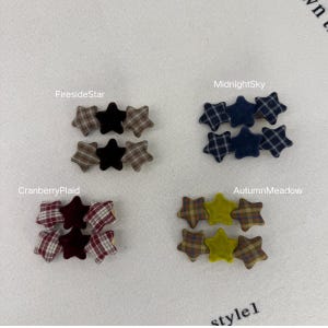 Puede incluir: Colección de adornos de tela en forma de estrella en varios colores y patrones. La imagen muestra conjuntos etiquetados como "FiresideStar", "MidnightSky", "CranberryPlaid" y "AutumnMeadow". Cada conjunto incluye tres estrellas, con diseños lisos y a cuadros.