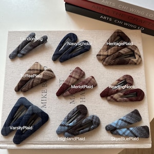 May include: A selection of plaid hair clips in shades of blue, brown, and navy. Each clip is labelled with a name like "LondonFog" and "NavyScholar". The clips are displayed on a neutral-coloured surface, with the text "ARTS CHI WING LO" visible.