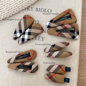 May include: Several hair clips in different shapes, adorned with a tan, red, black, and white plaid design. Styles include KensingtonClassic, SloaneTriangle, BelgraviaFrame, PrimrosePetite, and ChelseaBar. The background is a light beige fabric.