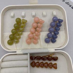 A flat lay photo showing four sets of beaded hair clips arranged on a white background.
Each set consists of two gold-tone alligator clips: one featuring a row of round spherical beads and the other featuring a row of heart-shaped beads.
The sets come in four glossy, jelly-like colors:

Toffee Brown (top left).
Peachy Pink (top right).
Matcha/Olive Green (bottom left).
Dusty Blue/Grey (bottom right).