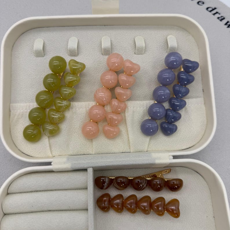 A flat lay photo showing four sets of beaded hair clips arranged on a white background.
Each set consists of two gold-tone alligator clips: one featuring a row of round spherical beads and the other featuring a row of heart-shaped beads.
The sets come in four glossy, jelly-like colors:

Toffee Brown (top left).
Peachy Pink (top right).
Matcha/Olive Green (bottom left).
Dusty Blue/Grey (bottom right).